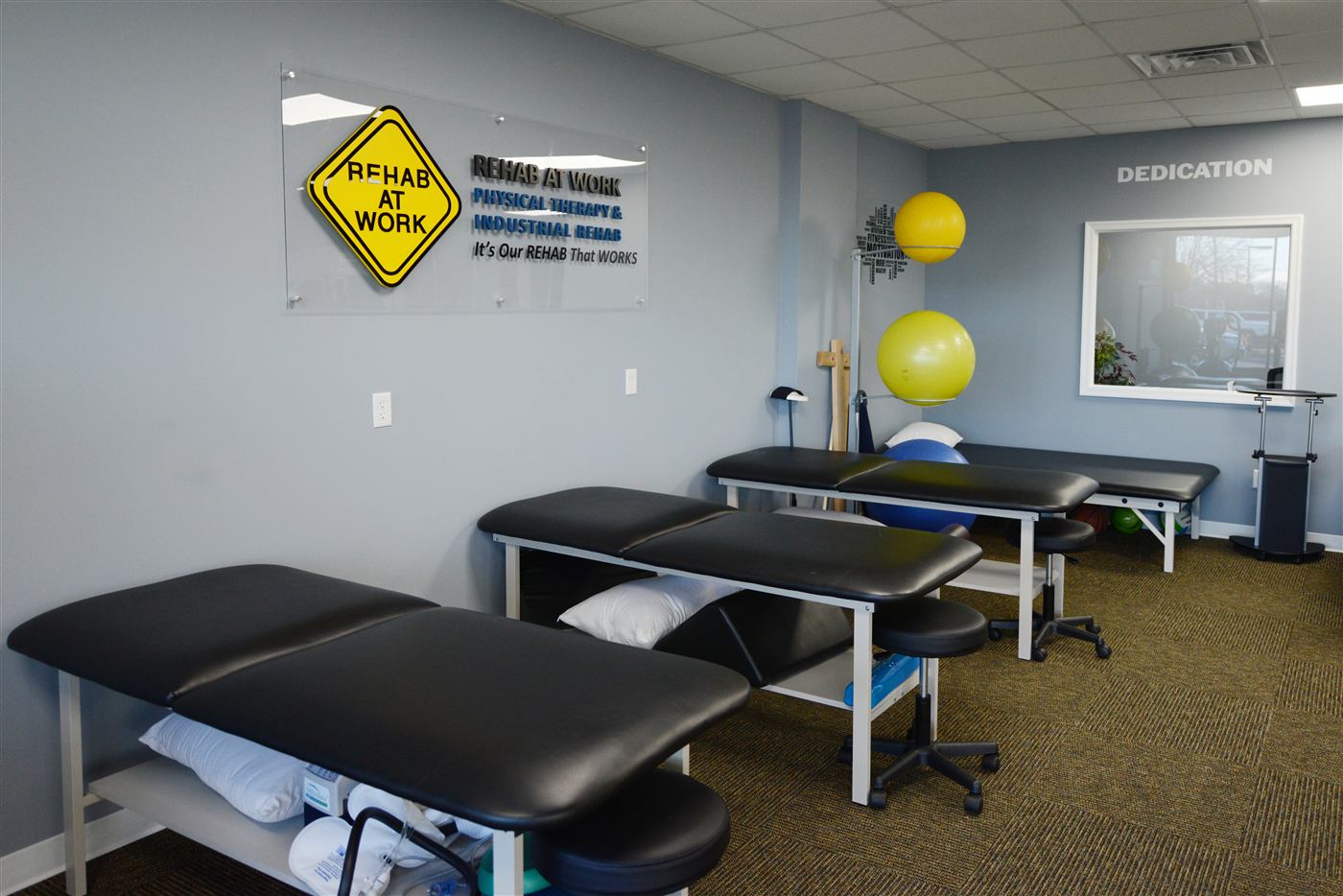 Clean treatment area with therapy tables, exercise balls and REHAB AT WORK signage
